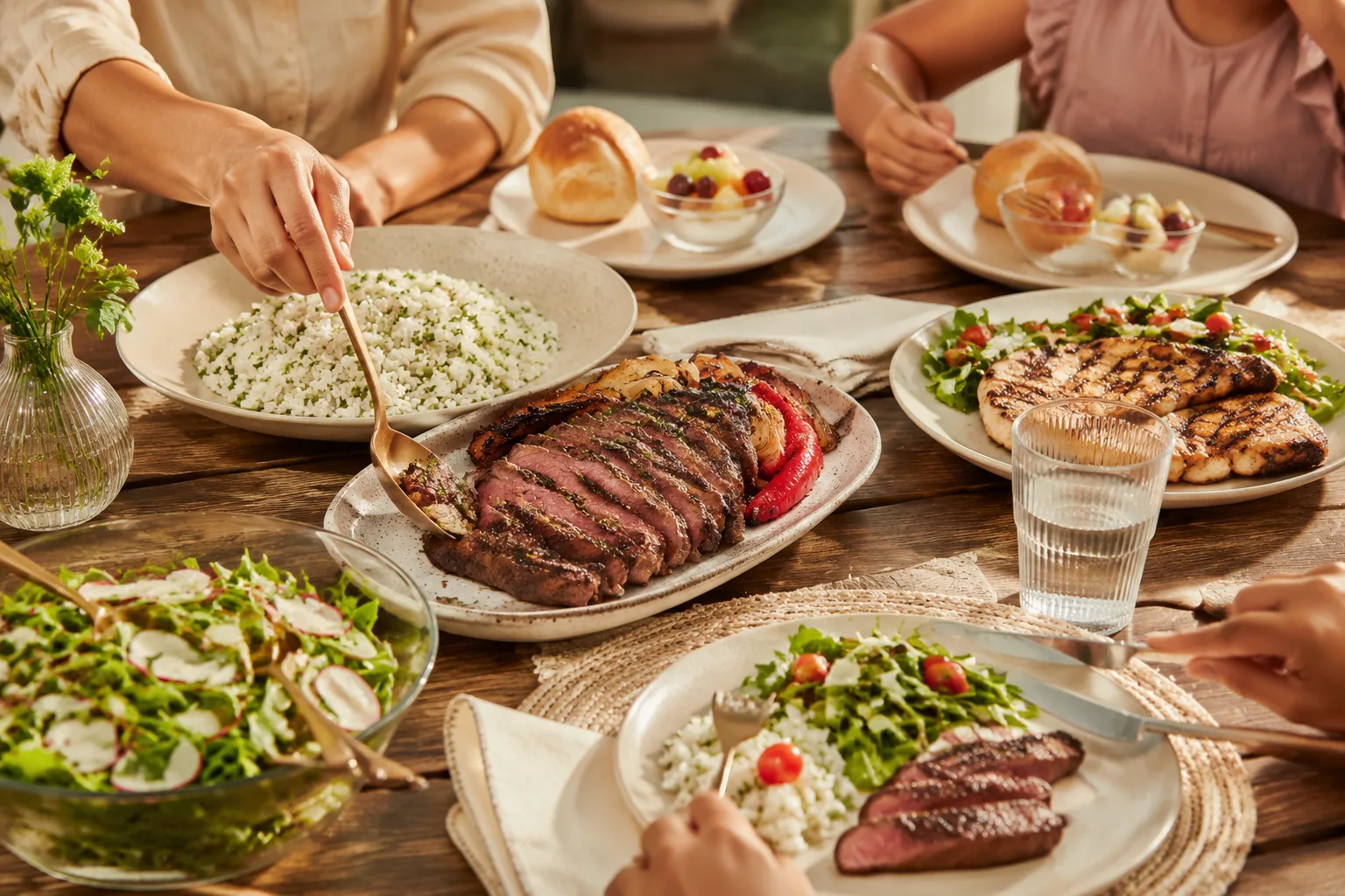 Family dinner table with several different plates being served, steak and rice, chicken and salad, and a smaller plate with a roll and fruit cup
